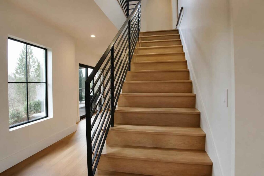 Drewniane schody prowadzące na piętro przy dużym oknie i czarnej metalowej balustradzie